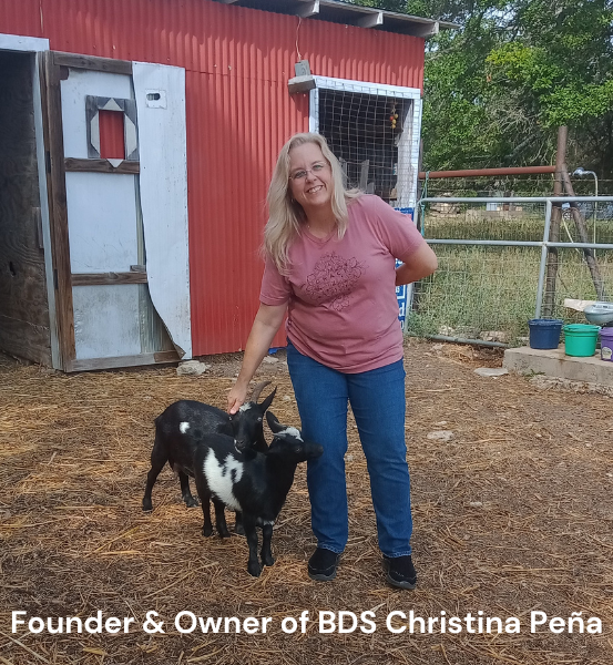 Woman standing with two baby goat's in front of a red barn, identified as the founder and owner of BDS Christina Peña.