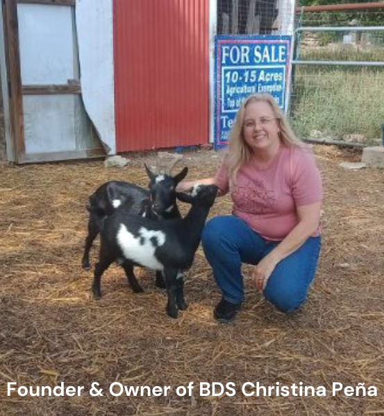 Woman kneeling next to two baby goat's in front of a red barn, identified as the founder and owner of BDS Christina Peña.