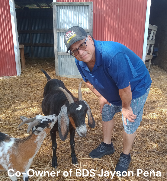Image of a Man standing next to two  baby goat's in front of a barn with the text Co-Owner of BDS Jayson Peña.